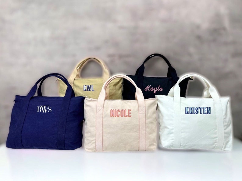 May include: Five personalized tote bags in various colors: navy blue, beige, black, cream, and off-white. Each bag features embroidered text in different colors, including initials and names. The bags have double handles and a rectangular shape.