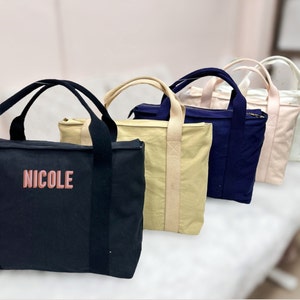 May include: A row of tote bags in various colors: black with "NICOLE" in pink, tan, navy blue, light pink, and white. Each bag has a rectangular shape, a zipper closure, and two handles.