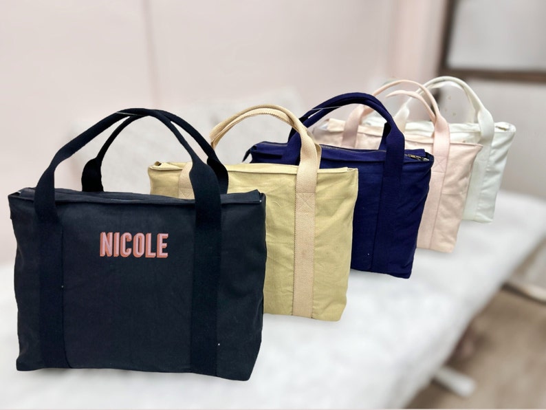 May include: A row of tote bags in various colors: black with "NICOLE" in pink, tan, navy blue, light pink, and white. Each bag has a rectangular shape, a zipper closure, and two handles.