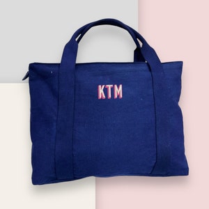 May include: A navy blue tote bag with two handles and a zipper closure. The bag features the embroidered letters "KTM" in white with a pink outline. The bag is made of a sturdy fabric and is designed for carrying items.