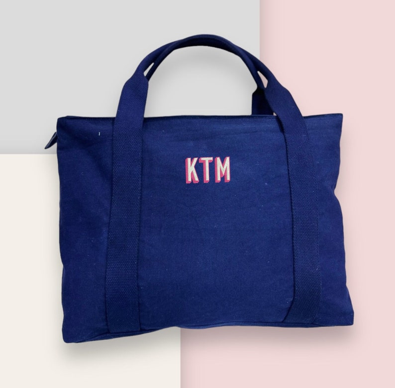 May include: A navy blue tote bag with two handles and a zipper closure. The bag features the embroidered letters "KTM" in white with a pink outline. The bag is made of a sturdy fabric and is designed for carrying items.