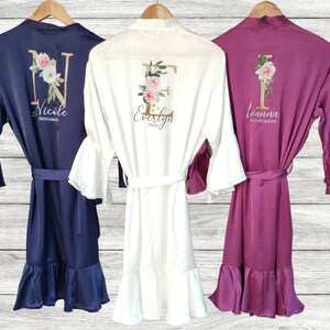 May include: Three satin robes with ruffled sleeves and hems. The robes are in navy blue, white, and burgundy. Each robe has a floral design with a gold initial and a name below. The names are "Nicole Bridesmaid", "Evelyn Bridesmaid", and "Ivanna Maid of Honor".