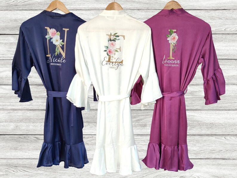 May include: Three satin robes with ruffled sleeves and hems. The robes are in navy blue, white, and burgundy. Each robe has a floral design with a gold initial and a name below. The names are "Nicole Bridesmaid", "Evelyn Bridesmaid", and "Ivanna Maid of Honor".