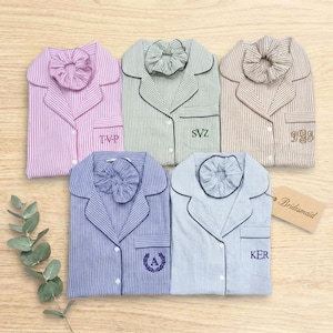 May include: Five button-down pajama shirts with matching scrunchies in pink, green, tan, purple, and blue stripes. Each shirt features a chest pocket with embroidered initials or monograms. A tan tag labeled Bridesmaid is attached to the blue shirt.