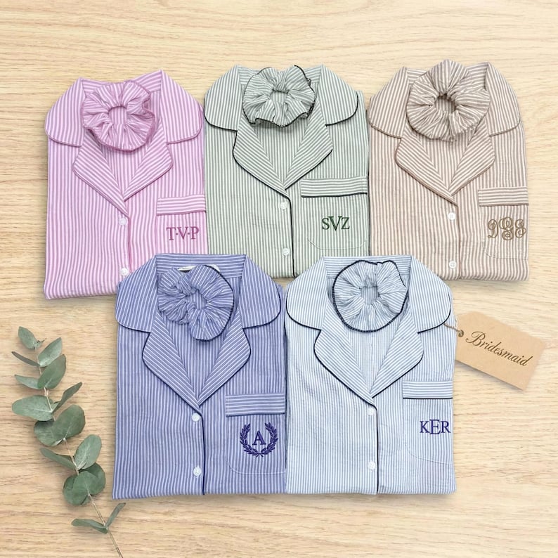 May include: Five button-down pajama shirts with matching scrunchies in pink, green, tan, purple, and blue stripes. Each shirt features a chest pocket with embroidered initials or monograms. A tan tag labeled Bridesmaid is attached to the blue shirt.