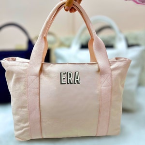 May include: A light pink tote bag with matching handles and trim. The bag features the letters "ERA" in a black and white block font on the front. Additional tote bags are visible in the background.