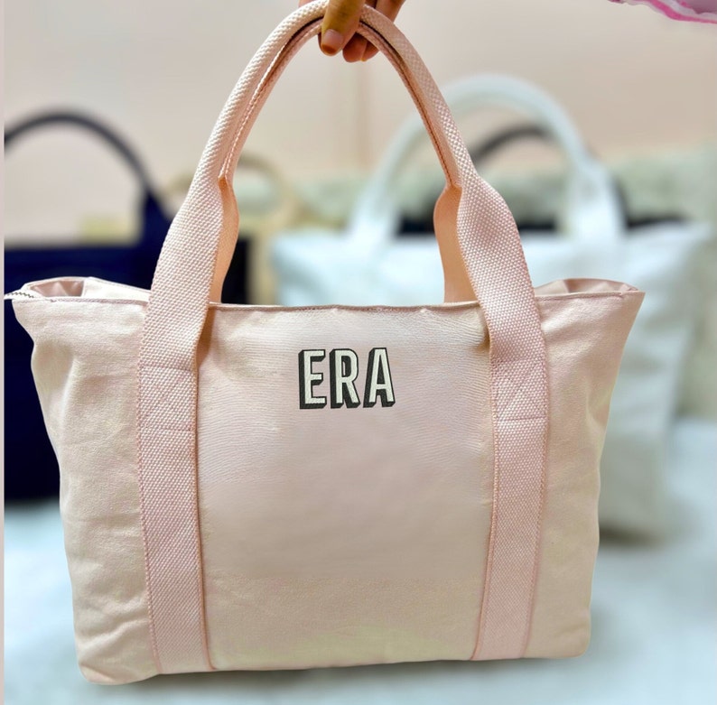 May include: A light pink tote bag with matching handles and trim. The bag features the letters "ERA" in a black and white block font on the front. Additional tote bags are visible in the background.