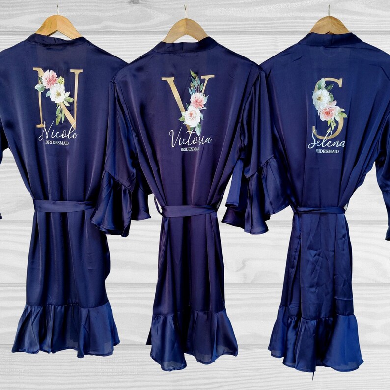 May include: Three navy blue satin robes with ruffled sleeves and hems. Each robe has a gold initial and floral design with the name "Nicole", "Victoria", and "Selena" embroidered on the back.