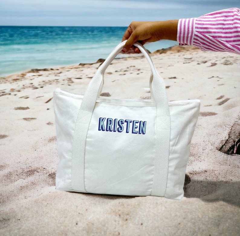 May include: White tote bag with the name "KRISTEN" in blue lettering. The bag has white handles and is held up against a beach background with the ocean in the distance.