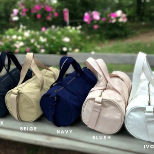 May include: Five canvas duffel bags in different colors: black, beige, navy, blush, and ivory. Each bag has a zipper closure and two top handles.