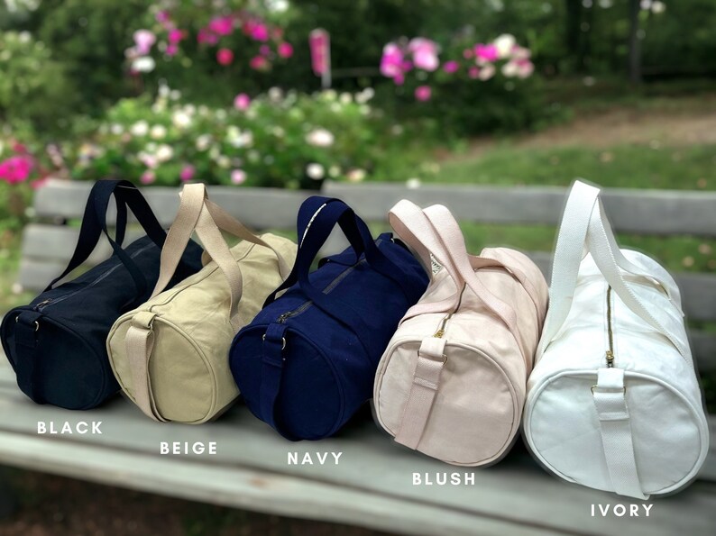 May include: Five canvas duffel bags in different colors: black, beige, navy, blush, and ivory. Each bag has a zipper closure and two top handles.