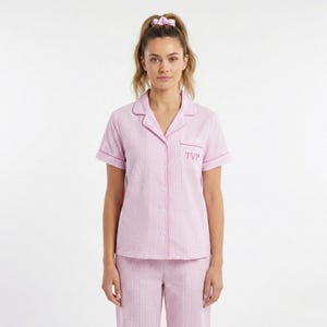 May include: Pink and white striped pajama set. The short-sleeved top has a button-down front, a breast pocket with the initials "TVP", and pink trim. The matching pants are also striped. A pink scrunchie is in the model's hair.