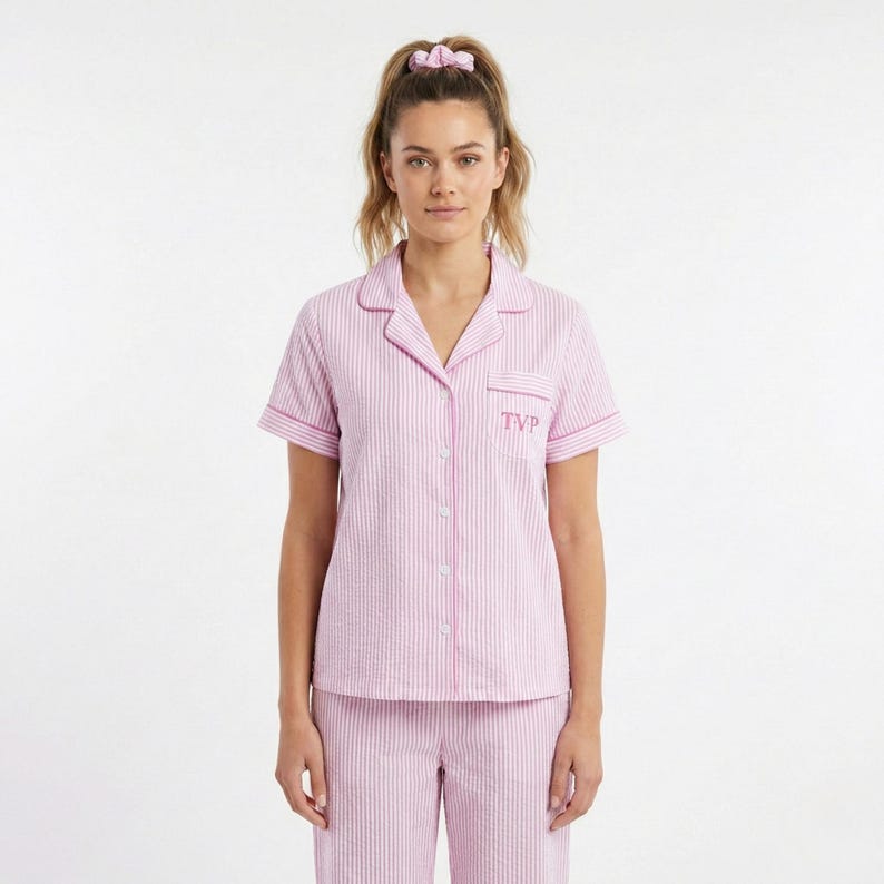 May include: Pink and white striped pajama set. The short-sleeved top has a button-down front, a breast pocket with the initials "TVP", and pink trim. The matching pants are also striped. A pink scrunchie is in the model's hair.