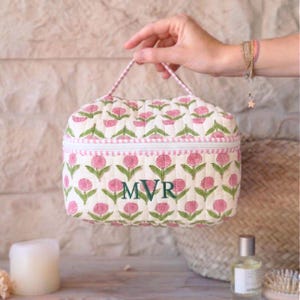 May include: A quilted cosmetic bag with a floral pattern in pink and green on a white background. The bag has a zipper closure and a handle. The initials "MVR" are embroidered in green on the front.
