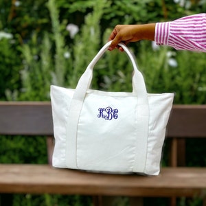 May include: A white canvas tote bag with matching handles and straps. The bag features a navy blue monogram in a script font. The bag is held up against a blurred green background.