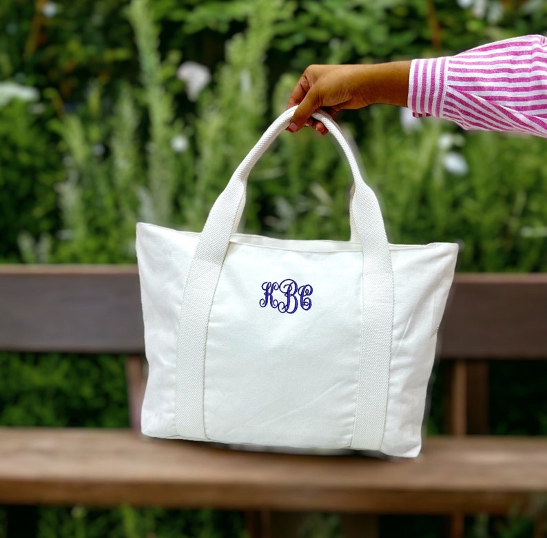May include: A white canvas tote bag with matching handles and straps. The bag features a navy blue monogram in a script font. The bag is held up against a blurred green background.