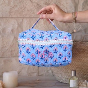 May include: A blue quilted cosmetic bag with a floral pattern in pink and blue. The bag has a zipper closure and a handle. The initials "KER" are embroidered in pink on the front.