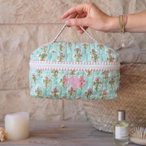 May include: A quilted cosmetic bag in a light blue floral pattern with pink accents and a pink monogram. The bag has a zipper closure and a handle. The bag is approximately 8 inches wide and 6 inches tall.