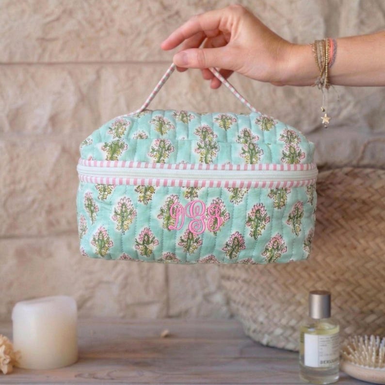 May include: A quilted cosmetic bag in a light blue floral pattern with pink accents and a pink monogram. The bag has a zipper closure and a handle. The bag is approximately 8 inches wide and 6 inches tall.