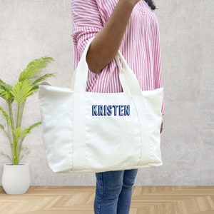 May include: A white tote bag with the name "KRISTEN" embroidered in blue and white. The bag has two white handles and is held by a person wearing a pink and white striped shirt and blue jeans.