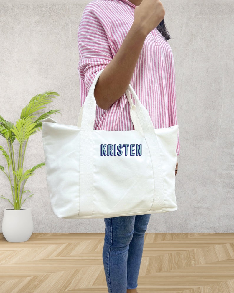 May include: A white tote bag with the name "KRISTEN" embroidered in blue and white. The bag has two white handles and is held by a person wearing a pink and white striped shirt and blue jeans.