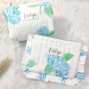 Puede incluir: Dos bolsas de maquillaje acolchadas blancas con estampado floral de hortensias azules y letras doradas. Las bolsas están personalizadas con el nombre "Evelyn".