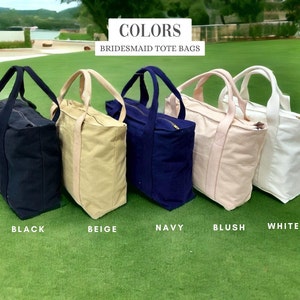 May include: Five canvas tote bags in black, beige, navy, blush, and white are displayed on green grass. Each bag has a matching handle and a zipper closure. The text "COLORS BRIDESMAID TOTE BAGS" is at the top of the image.