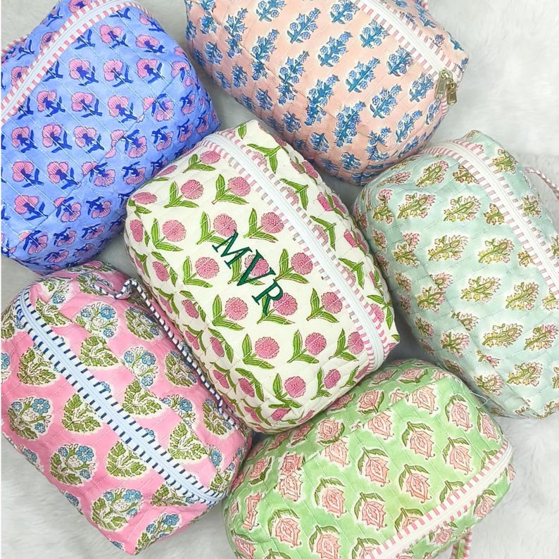 May include: A collection of quilted cosmetic bags in various colors, including blue, pink, and green. Each bag features a floral pattern and a zipper closure. One bag has the initials "MVR" embroidered on it.