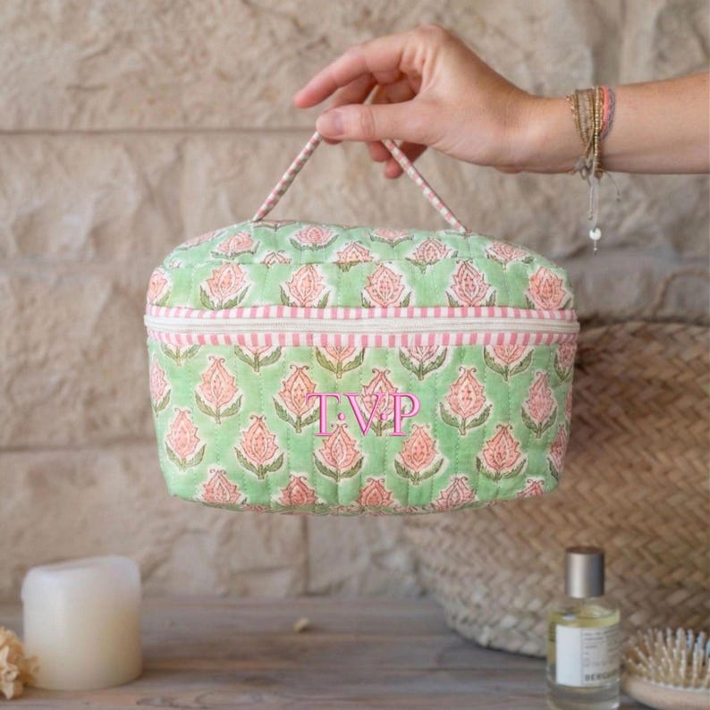 May include: A quilted cosmetic bag with a green background and a repeating pink floral pattern. The bag has a pink striped border and a handle. The initials "TVP" are embroidered on the front.