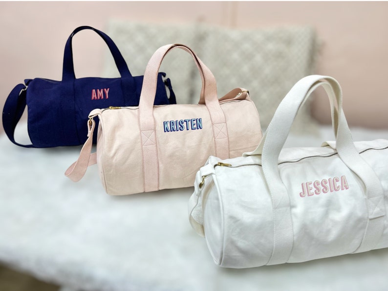 May include: Three personalized canvas duffel bags in navy blue, pink, and white. The bags have the names "Amy", "Kristen", and "Jessica" embroidered on them in different colored thread.