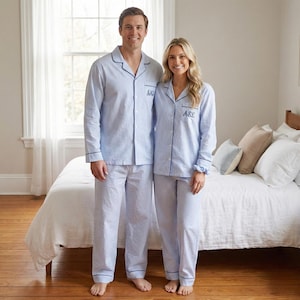 May include: Matching light blue and white striped pajamas with navy blue trim. The pajamas have a button-down shirt and pants. The shirts are monogrammed with initials in navy blue. The couple is standing in a bedroom.