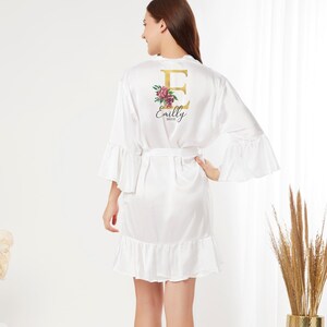 May include: White satin robe with a ruffled hem and bell sleeves. The robe has a gold letter "E" with floral accents and the name "Emily Bride" printed on the back.
