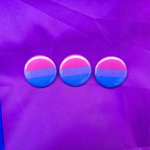 May include: Three round, colorful buttons with the bisexual pride flag colors: pink, blue, and purple. The buttons are on a purple background.