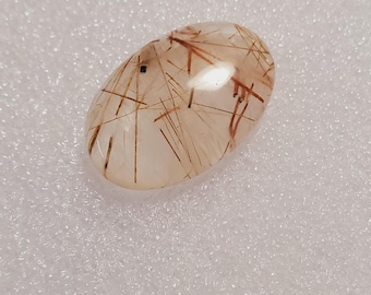 May include: Oval-shaped gemstone with a clear base and intricate brown needle-like inclusions. The polished stone has a smooth surface and is set against a white background. A unique and natural crystal.
