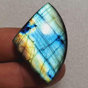 May include: A polished, iridescent labradorite gemstone with a unique shape. The stone displays a vibrant play of color, with blues, yellows, and hints of green. The surface has a grid-like pattern, enhancing its natural beauty. This gemstone is ideal for jewelry making.