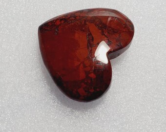May include: A heart-shaped stone with a deep red color and dark brown marbling. The polished surface reflects light, and the stone is set against a white background. A unique decorative item.