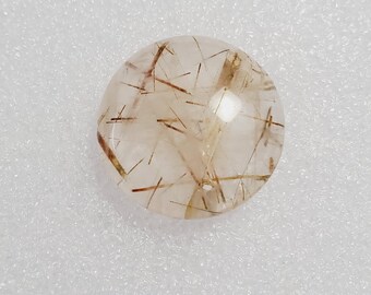 May include: A clear, round gemstone with internal golden rutile inclusions. The gemstone has a polished surface and is set against a white background. The inclusions create a striking pattern.