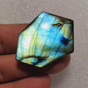 May include: A polished labradorite gemstone, cut into an unusual shape. The stone exhibits a shimmering display of colour, with blues, greens, and yellows blending across its surface. Darker markings add depth to the stone's appearance.