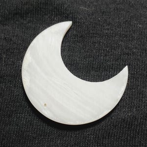 Può includere: Un oggetto bianco a forma di mezzaluna. La luna ha una superficie liscia, leggermente strutturata e una forma arrotondata. L'oggetto è probabilmente un oggetto decorativo o un gioiello, forse fatto di pietra o conchiglia.