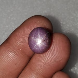 May include: A round, polished, purplish-red gemstone with a six-rayed star pattern. The star effect, or asterism, is visible across the surface of the gem. The stone is held between fingers, showcasing its colour and shape.