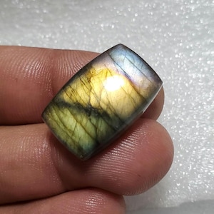 Può includere: Una pietra preziosa di labradorite rettangolare con una superficie lucida. La pietra mostra un mix di colori, tra cui giallo, verde, blu e viola, con un effetto scintillante. La pietra preziosa è tenuta tra le dita.