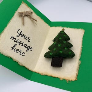 May include: A handmade Christmas card with a green felt Christmas tree and the text "Your message here". The card is open, revealing a cream-colored paper with a torn edge. A small bow is attached to the top.