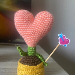 May include: A crocheted heart-shaped plant with green leaves and a brown pot. The pot is yellow and has a crocheted texture. A pink heart with an arrow is attached to a wooden stick.