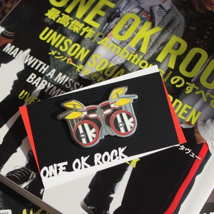 Könnte beinhalten: Eine silberne und schwarze Schlagzeug-Anstecknadel mit gelben Becken. Die Nadel trägt die Aufschrift "ONE OK ROCK" in weiß auf schwarzem Hintergrund.