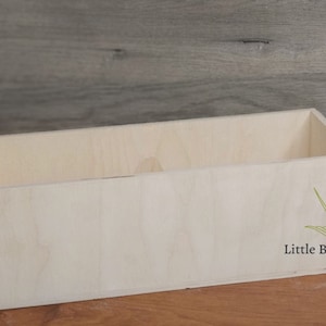 May include: A rectangular, unfinished wooden box with a light, natural wood grain. The box is open at the top and has a simple, clean design. A green illustration of a bird and the text "Little Bird Shop" are on the side.