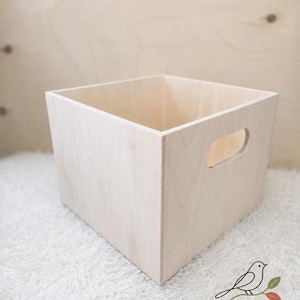 May include: A square, light-colored wooden storage box with a smooth finish and a cutout handle on the side. The box features clean, straight edges and an open top. In the bottom right corner, there is a logo with a bird and the text Little Bird SHOP STUDIO Little wonders, made for big smiles.