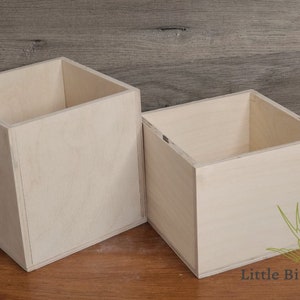 May include: Two unfinished wooden square boxes. The boxes are made of light-colored wood and have an open top. The boxes are sitting on a wooden surface. The boxes are likely for storage or display.