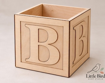 May include: A square wooden box featuring a large, laser-cut letter B on the front and side panels. The light-colored wood has dark, defined edges, and the box is open at the top. The logo Little Bird Shop Studio is visible in the corner.