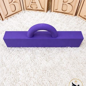 May include: A solid purple rectangular block with a matching purple arched handle attached to the top center. The object is positioned in front of three light-colored wooden blocks that each feature a single embossed letter, spelling out BAB.