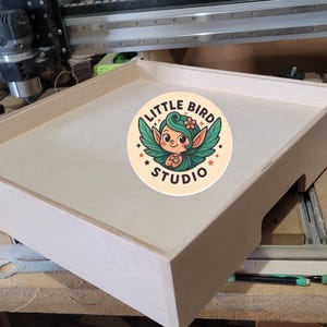 May include: A light-colored wooden tray with a recessed interior and a cutout on one side. The tray features a circular sticker with a cartoon fairy and the text "LITTLE BIRD STUDIO". The tray is likely for crafting or storage.